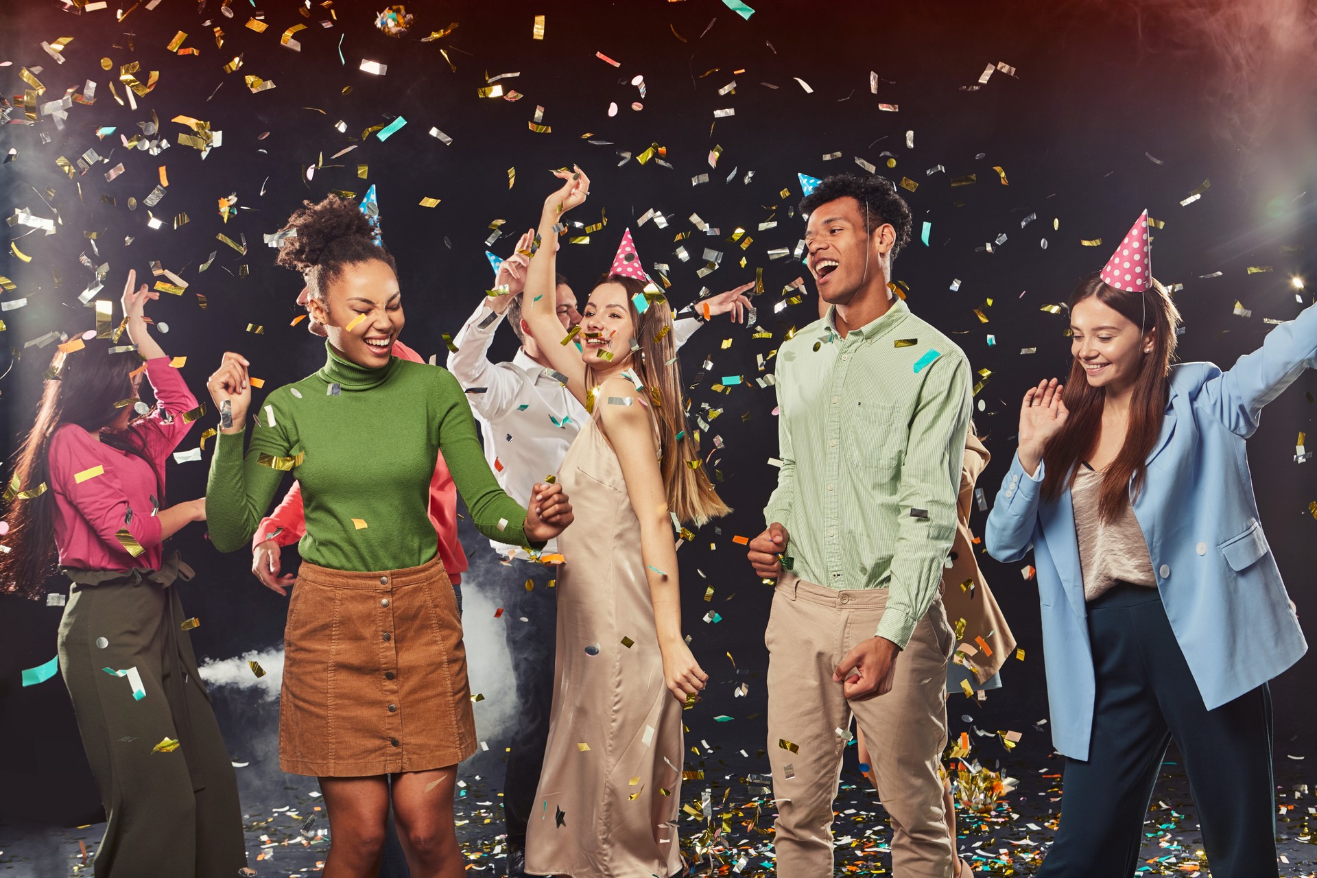 Group of young happy multiracial people wearing birthday hats dancing and having fun, confetti falling in the air